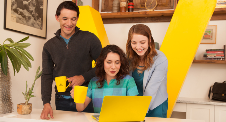 man-and-woman-standing-behind-woman-sitting-with-yellow-laptop-computer-and-yellow-coffee-mugs-with-yellow-Vantage-West-V-in-background