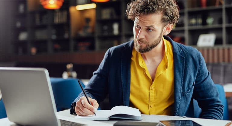 man-looking-at-computer-taking-notes-with-pen-and-notebook