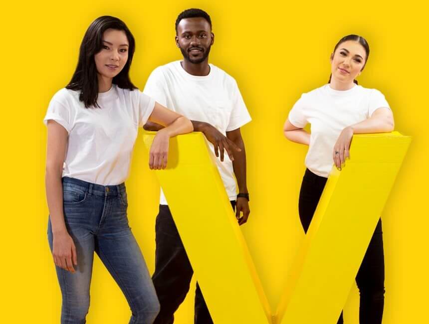 Three students in white t-shits stand in behind the yellow Vantage West "V."